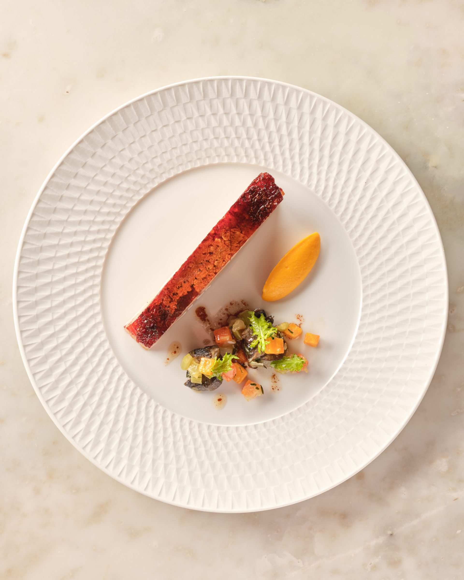 Gourmet dessert with a long red layered slice, orange quenelle and diced fruit garnish on a textured white plate, top view.