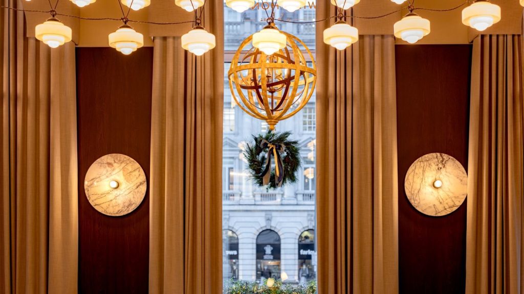 The interior of Wild Honey St James dressed for Christmas Eve, with a festive wreath in the window, golden globe chandelier and warm ambient lighting overlooking Pall Mall